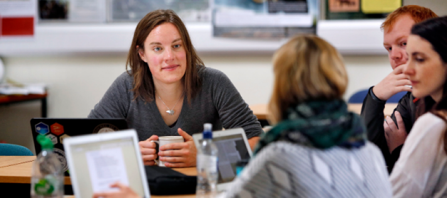 A woman holding a coffee cup seated at a desk surrounded by colleagues and laptops