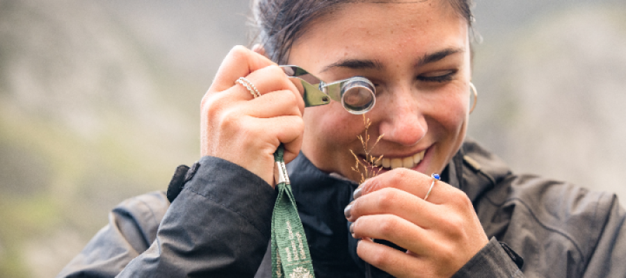 A female student outdoors in the forest using a looking glass to inspect a plant sample