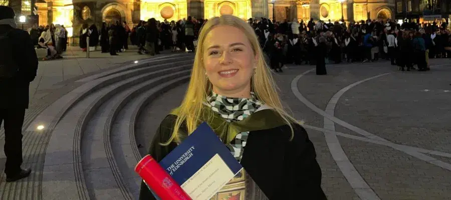 Anna wearing her graduation gown and holding her degree scroll, standing in front of McEwan Hall