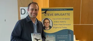 Professor Steve Brusatte holding up his book, The Story of Birds, in front of a poster with further information about it.