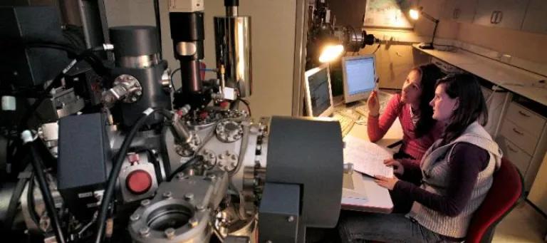 Two female scientists seated looking at computer monitors and surrounded by scientific equipment including Ion Micro-Probe