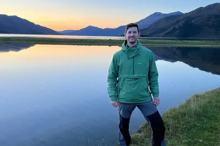 Ross stands in front of a lake with mountains in the background