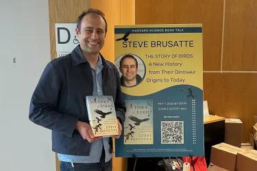 Professor Steve Brusatte holding up his book, The Story of Birds, in front of a poster with further information about it.
