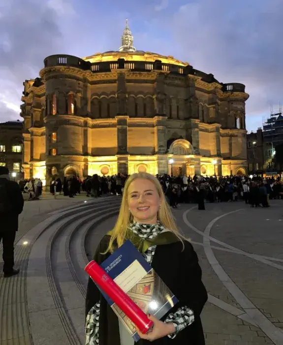 Anna at her graduation, holding her degree certificate and scroll in front of McEwan Hall