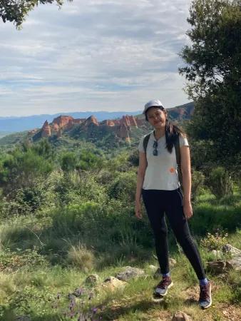 Via smiles for the camera, standing in front of a mountainous landscape filled with greenery such as plants and trees.