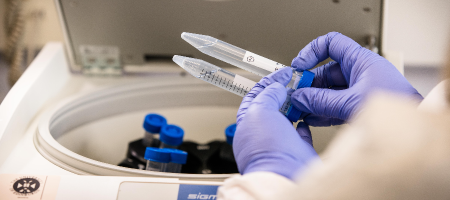 Blue gloved hands holding a dropper in front of a scientific centrifuge filled with test tubes