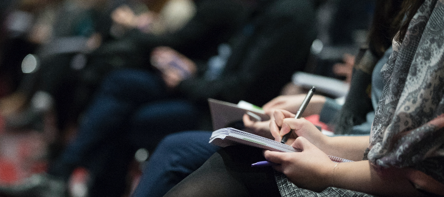 A woman's hands writing in a notebook on her lap, seated next to a group of people at a lecture