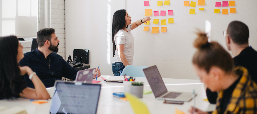 A woman standing and pointing to post-it notes on a board, while a table of colleagues with laptops observe