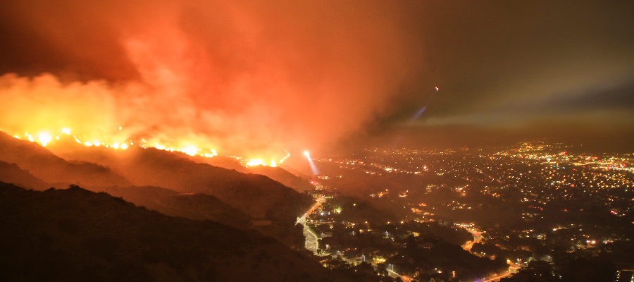 Wildfire on hills burning towards city suburbs at night, with helicopter and rescue teams evacuating neighborhood