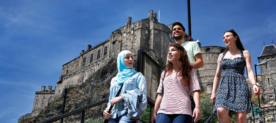 One male and three female students outdoors walking down steps with Edinburgh castle in the background