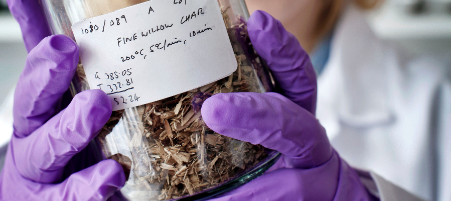 Surgical gloved hands holding a jar containing biochar. The jar label says 'willow biochar'